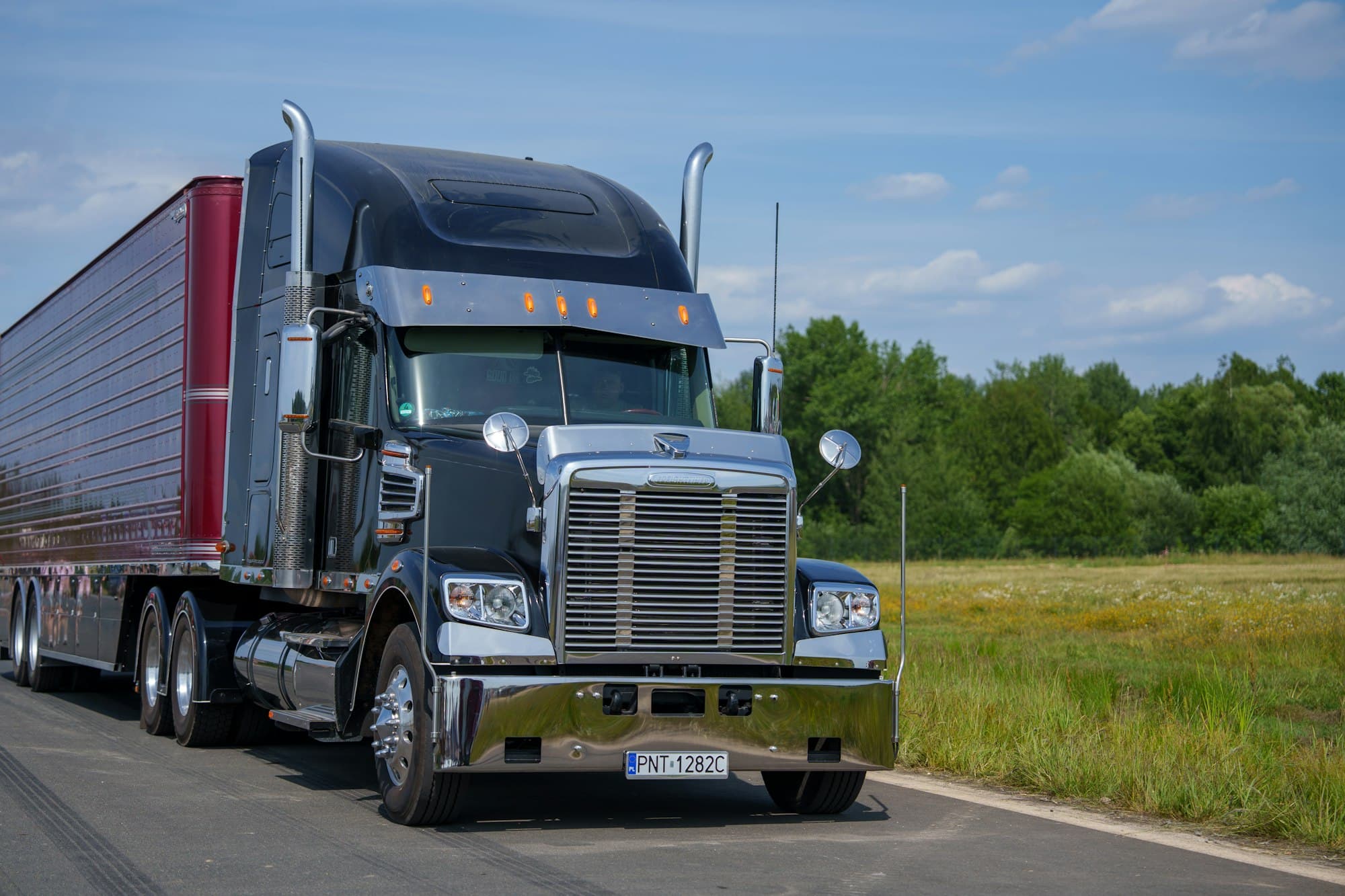 A dark tractor pulling a dry van past a green field